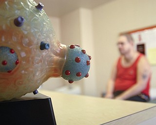 Michael Bushara, a gay, white male from Youngstown, discusses possible reasons for his lowered T-cell count with a doctor at an HIV/AIDS clinic. In the foreground is a model of dark blue HIV viruses replicating within a healthy human T-cell.