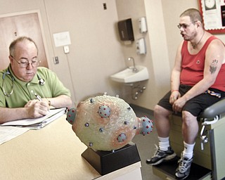 Michael Bushara, a gay, white male from Youngstown, discusses possible reasons for his lowered T-cell count with a doctor at an HIV/AIDS clinic. In the foreground is a model of dark blue HIV viruses replicating within a healthy human T-cell.