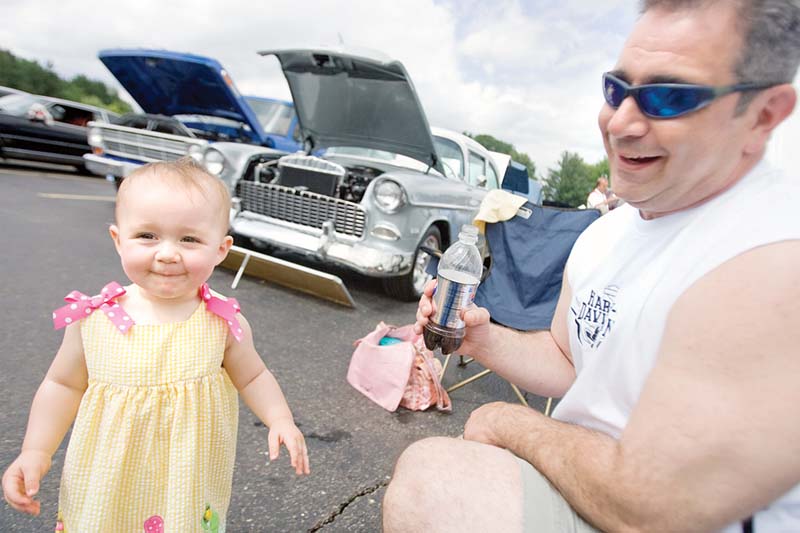 13 month old Kalli Stephens, of Ravenna, smiles after her taking a drink of her father, John Stephens', beverage during the annual Fathers Day Car Show at Springfield High School on Sunday afternoon. John's stepfather's 1955 Silver Cheverolet can be seen in the background and to the left of that sits John's 1967 Blue Ford Truck.