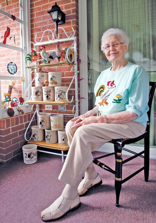 FAIR COLLECTOR:  Eileen Groves, a Canfield native, sits in her home in front of a collection of 13 commemorative crocks featuring the Canfield Fair rooster mascot and slogan, “Something to crow about.” Groves now resides at Copeland Oaks Retirement Community in Sebring.