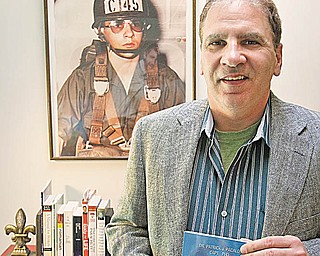 Patrick Pacalo stands in his Boardman home holding a copy of his latest book, “Eastern Europe: Cold Warfare III.” The photo on the wall is of Pacalo at age 17 in the Pa. Army National Guard.