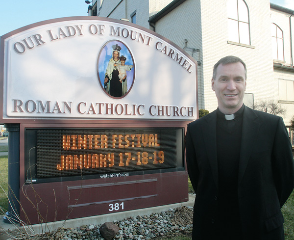 The Rev. John-Michael Lavelle, pastor of Our Lady of Mount Carmel Church, Niles, stands next to a sign promoting the winter festival this weekend. The church’s summer festival, a tradition for more than 80 years, inspired the January event to satisfy those summer food cravings.