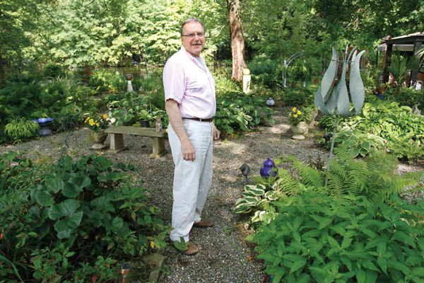 The Rev. David W. Merzweiler, left, better known as Pastor Dave, stands on a gravel path in the expansive garden he has worked on behind the rectory at St. Catherine Church, 1254 Grandview Road, Lake Milton, where he lives.
The green oasis of plants also features garden art, statues, four fountains, a pond, wind chimes and an enclosure
with four peacocks. 