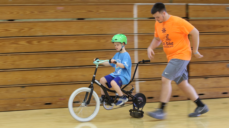 Seth Baker rides a specially adapted bike while Ryan Jones jogs to keep up at the iCAN Shine Bike Camp at Boardman Glenwood Junior High School. The camp continues through Friday.